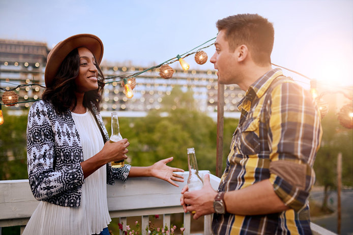 A woman in a hat and a man in a plaid shirt talk on a balcony at dusk overlooking a city. They are holding beverages and a string of outdoor lights hangs in the background.